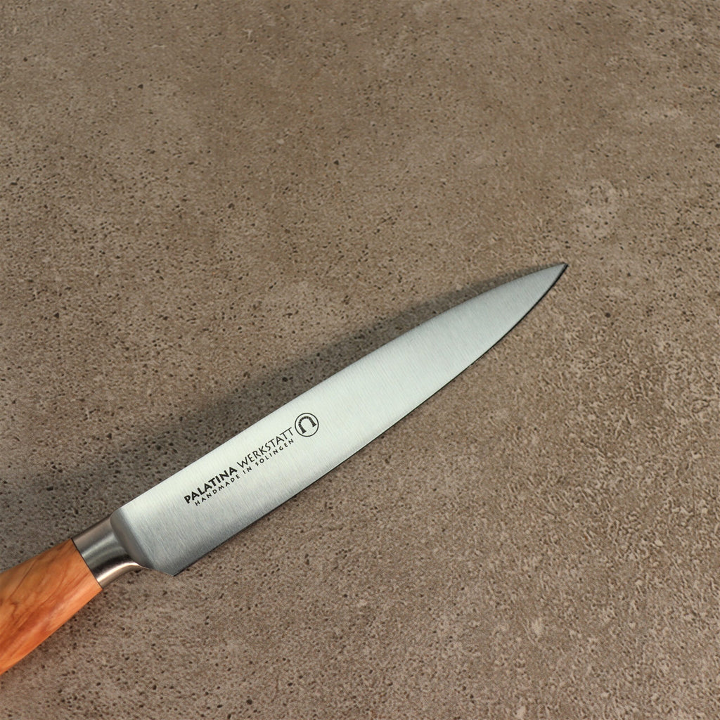 Ein 12 cm langes Officemesser der Palatina Werkstatt Messerserie mit Olivenholzgriff liegt auf einer strukturierten grauen Oberfläche. Auf der Klinge sind die Inschriften „PALATINA WERKSTATT“ und „HANDMADE IN GERMANY“ sowie ein kleines rundes Logo zu sehen.