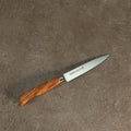 Das Palatina Werkstatt Messerserie Officemesser 12 cm mit Olivenholzgriff und scharfer, spitzer Klinge aus rostfreiem Stahl liegt auf einer strukturierten grauen Oberfläche. In der Nähe des Griffs der Klinge ist ein Text eingraviert, der auf die Handwerkskunst der Palatina Werkstatt hinweist.