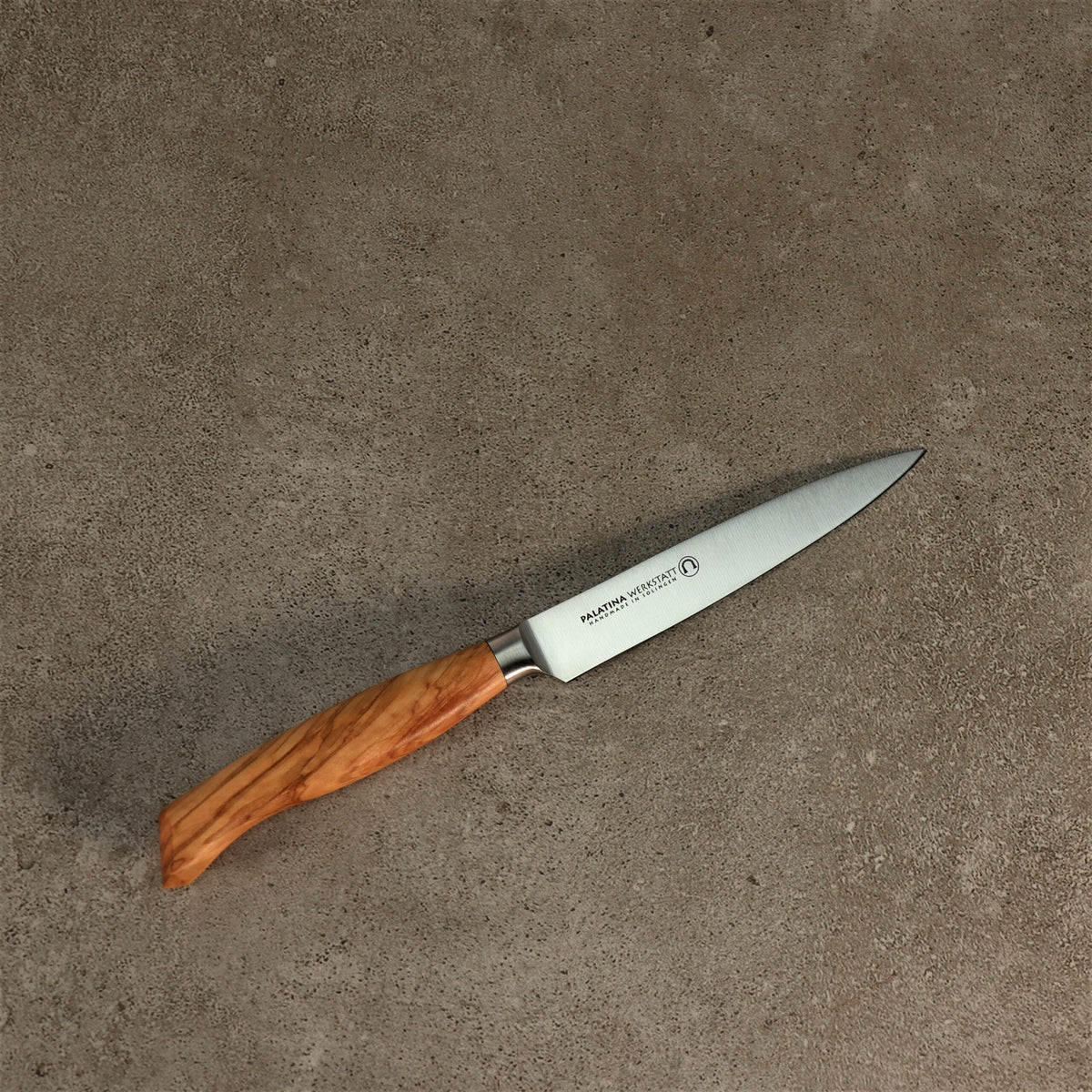 Das Palatina Werkstatt Messerserie Officemesser 12 cm mit Olivenholzgriff und scharfer, spitzer Klinge aus rostfreiem Stahl liegt auf einer strukturierten grauen Oberfläche. In der Nähe des Griffs der Klinge ist ein Text eingraviert, der auf die Handwerkskunst der Palatina Werkstatt hinweist.