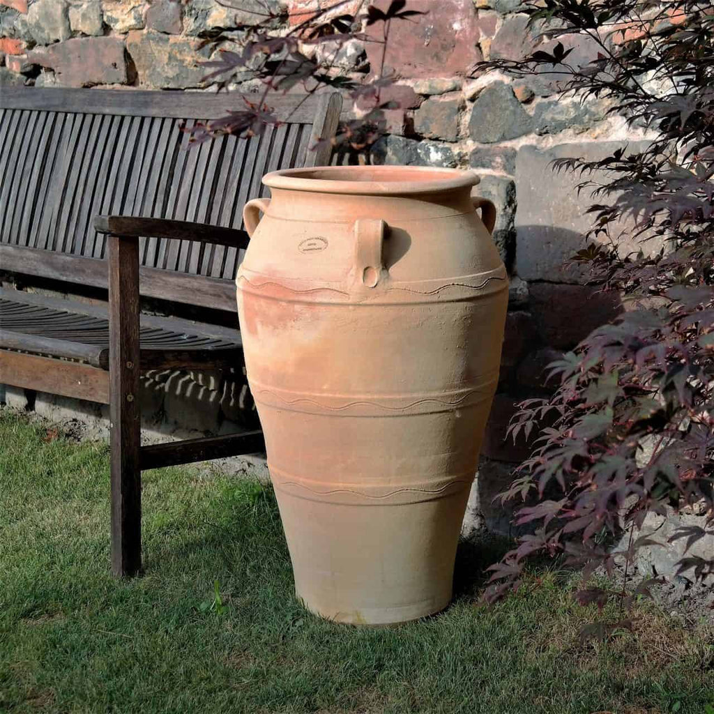 Eine große, frostfeste Terrakotta-Amphore für den Garten „Olea“ H=70 cm steht auf Gras neben einer Holzbank, dahinter eine Steinmauer.