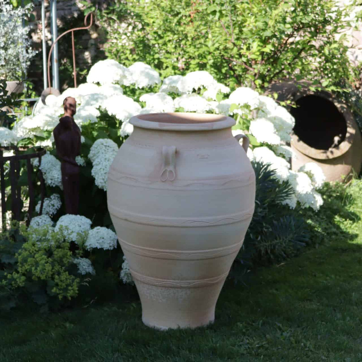 Eine frostfeste Terrakotta-Amphore „Olea“ (H= 100 cm) steht in einem üppigen Garten, umgeben von blühenden weißen Hortensien. Im Hintergrund sind eine kleine Statue und andere Gartendekorationen, darunter ein Olea-Baum, zu sehen, die die ruhige und natürliche Umgebung noch verstärken.