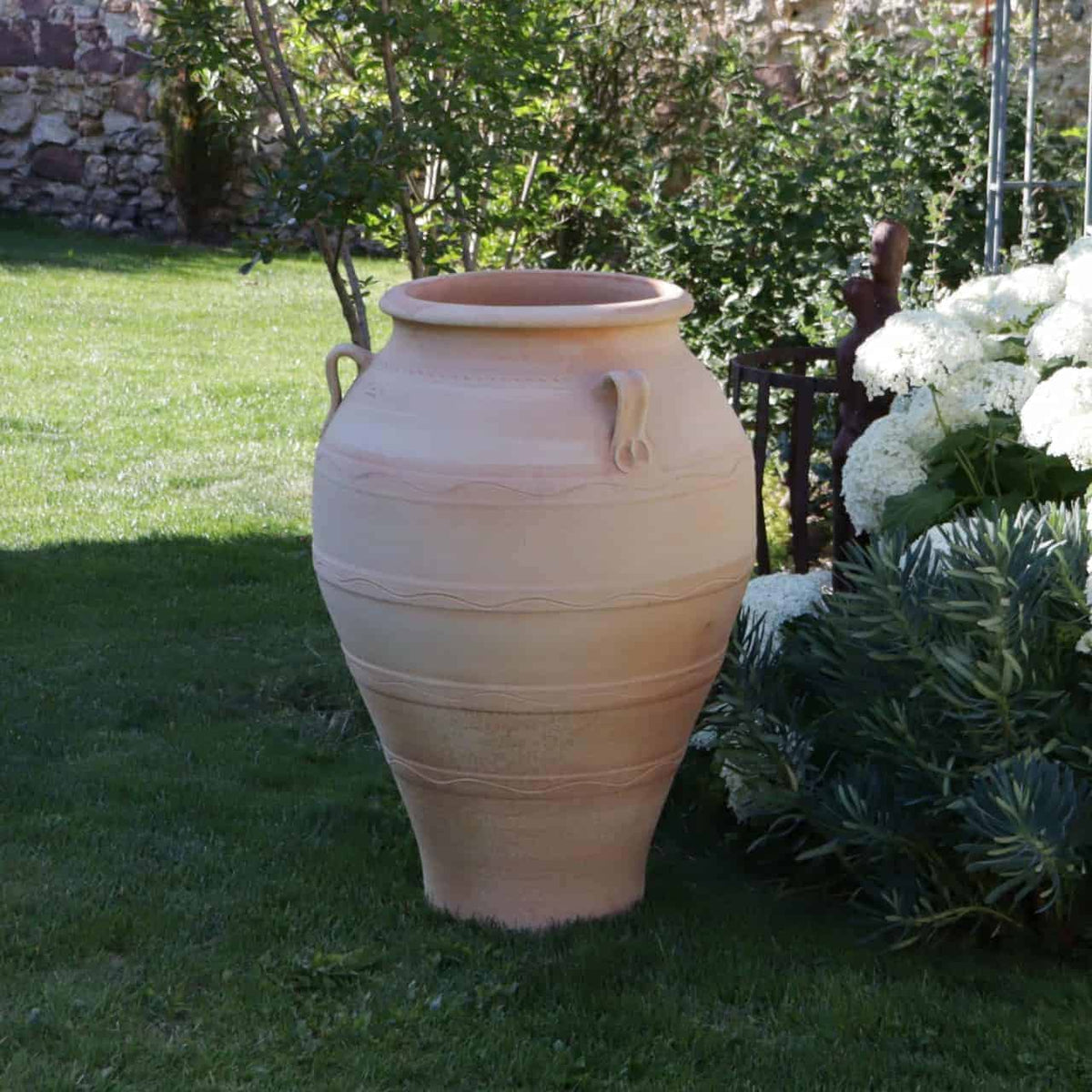 Eine große, beige frostfeste Terrakotta-Amphore „Olea“ (Höhe 100 cm) mit zwei Henkeln steht aufrecht auf einem gepflegten Rasen. Sie ist von üppigem grünem Laub und blühenden weißen Blumen umgeben, im Hintergrund ist teilweise eine Steinmauer zu sehen. Die Szene wird durch einen in der Nähe gepflanzten Olea-Baum noch verstärkt.