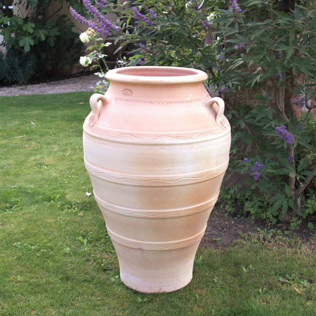Eine große, frostfeste Terrakotta-Amphore „Olea“ in Beige, 100 cm hoch, mit dekorativen Henkeln und aufwendigen Mustern, steht auf saftig grünem Gras, umgeben von einem Garten mit Blattpflanzen und violett blühenden Büschen.