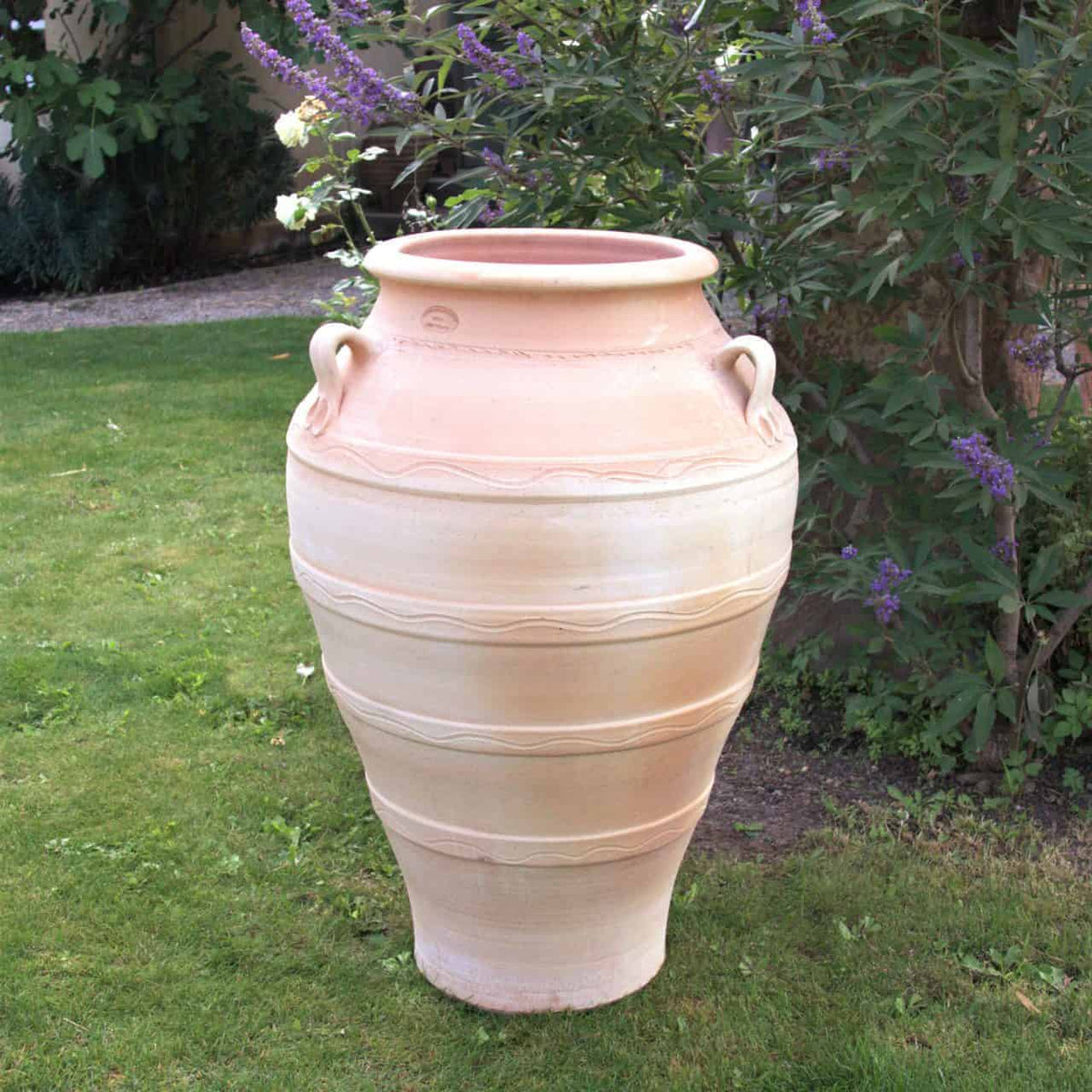 Eine große, frostfeste Terrakotta-Amphore „Olea“ in Beige, 100 cm hoch, mit dekorativen Henkeln und aufwendigen Mustern, steht auf saftig grünem Gras, umgeben von einem Garten mit Blattpflanzen und violett blühenden Büschen.