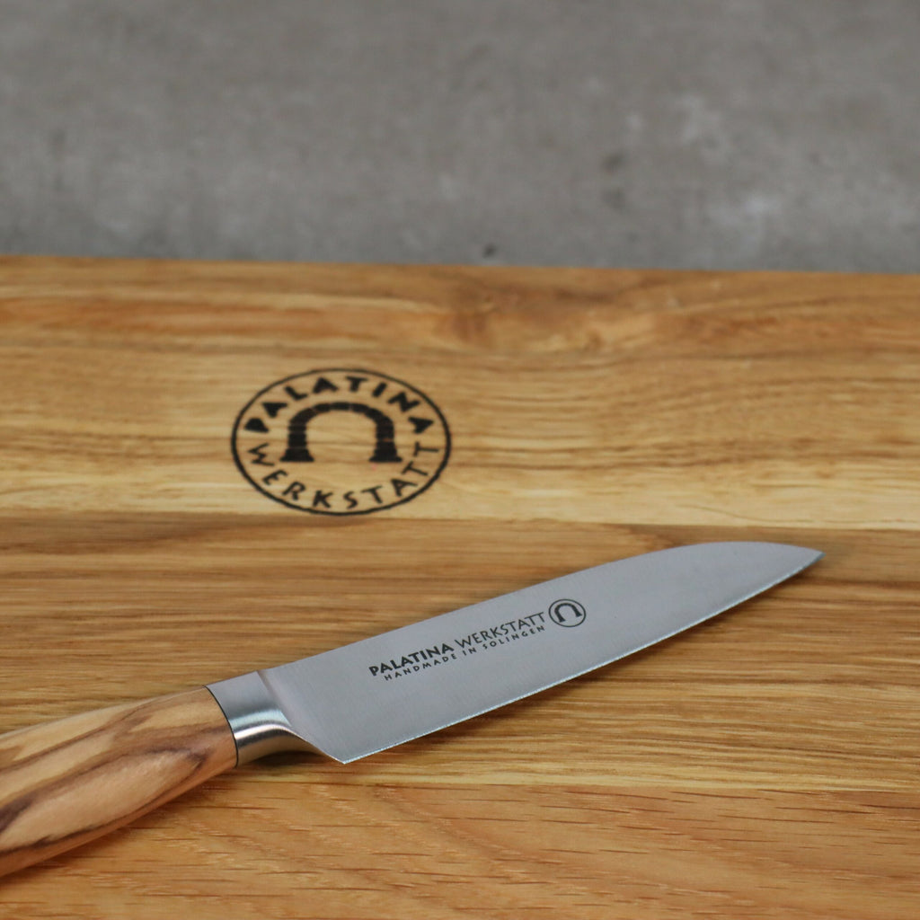 Das „Palatina Werkstatt Messerserie kleines Gemüsemesser“ mit 9 cm langer Klinge und Olivenholzgriff ruht auf einem Holzschneidebrett. Das Schneidebrett ist mit einem eingravierten Logo der „Palatina Werkstatt“ versehen, das ein Bogensymbol und den Text „Handmade in Germany“ enthält.