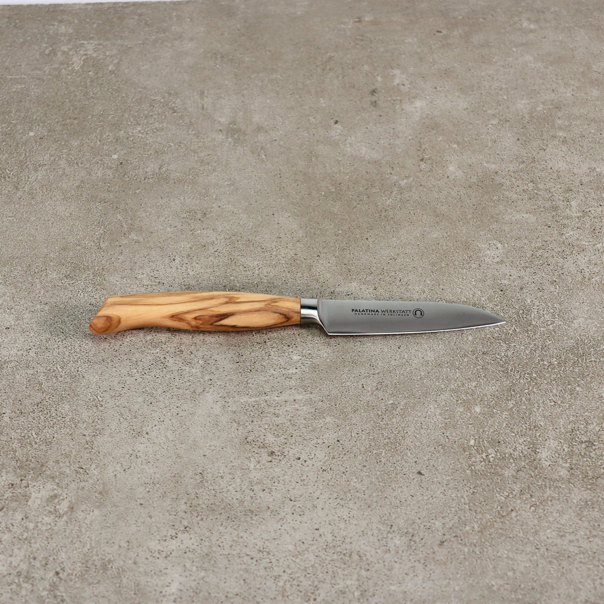 Ein kleines Gemüsemesser der Palatina Werkstatt Messerserie mit 9 cm langer Klinge und Olivenholzgriff liegt auf einer strukturierten grauen Oberfläche. In die Klinge aus rostfreiem Stahl sind neben dem Griff ein Text und ein Logo eingraviert.