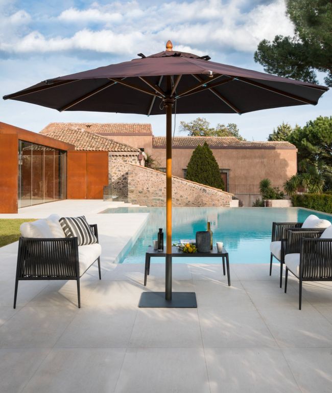 Eine luxuriöse Außenszene mit einem Swimmingpool, der von modernen Sitzgelegenheiten umgeben ist. Zwei schwarz-weiße gepolsterte Stühle stehen auf beiden Seiten eines kleinen Tisches unter einem großen, dunklen Runder Sonnenschirm Lipari 350 - 400 cm. Im Hintergrund sind Steingebäude mit Ziegeldächern und üppigem Grün zu sehen, die an Lipari erinnern.