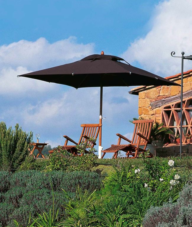 Eine ruhige Außenterrasse mit Holzstühlen und einem Tisch unter dem großen quadratischen Sonnenschirm Lipari, der 300 - 400 cm breit ist und reichlich Schatten spendet. Der Bereich ist mit üppigem Grün und blühenden Blumen geschmückt, vor der Kulisse eines Steingebäudes und eines teilweise bewölkten blauen Himmels.