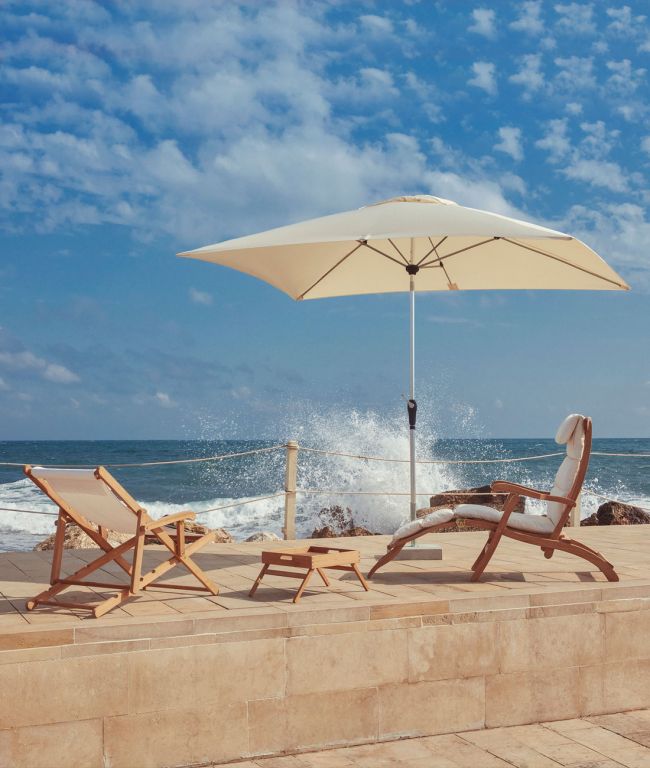 Eine ruhige Strandszene mit hölzernen Liegestühlen und einem kleinen Tisch unter einem verstellbaren rechteckigen Sonnenschirm Lipari mit Aluminium. Wellen schlagen gegen das Ufer unter einem strahlend blauen Himmel mit vereinzelten Wolken. Die Umgebung ist ruhig, ideal zum Entspannen.