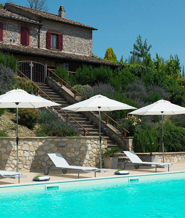 Ein wunderschönes Steinhaus mit roten Fensterläden liegt auf einem üppig grünen Hügel und bietet Blick auf einen glitzernden Swimmingpool. Weiße Liegestühle und verstellbare Lipari-Sonnenschirme mit Aluminiumstangen säumen den Pool und schaffen einen ruhigen und einladenden Außenbereich unter einem klaren blauen Himmel.
