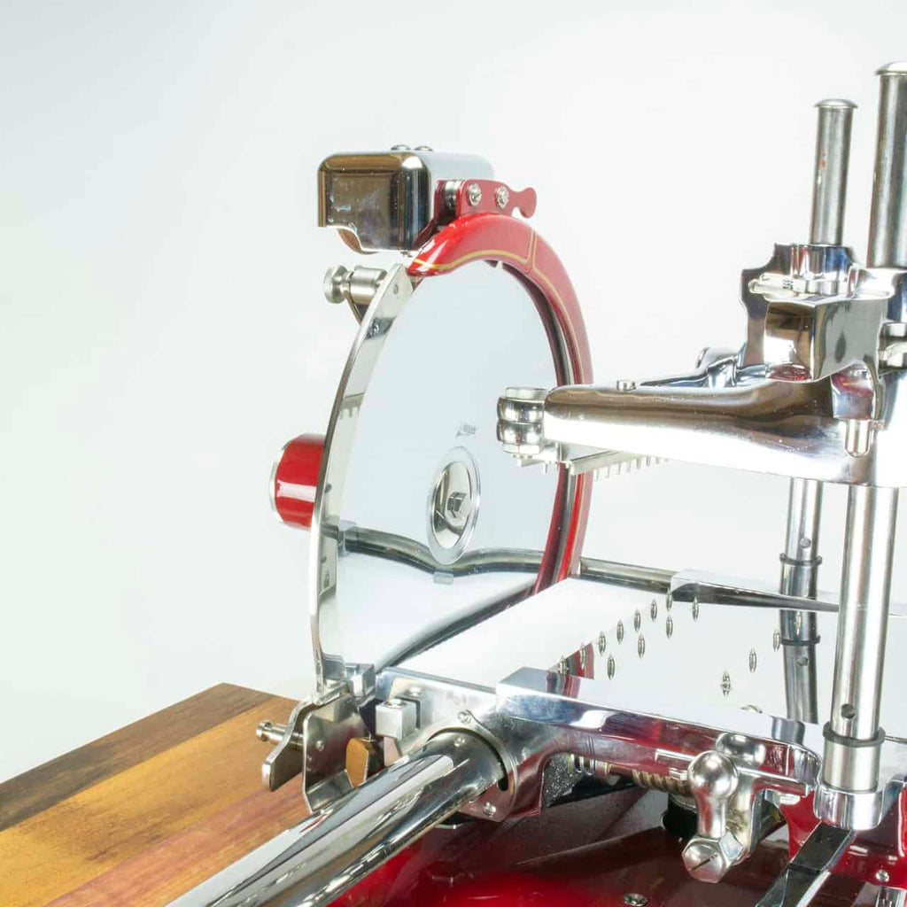 Eine Nahaufnahme einer Berkel Volano P15 Rot, Aufschnittmaschine Schwungrad + Standfuß + Aufschnittbrett, in Rot und poliertem Chrom. Diese Schwungrad-Aufschnittmaschine verfügt über ein großes rundes Messer, Einstellknöpfe und eine Holzplattform. Die glänzenden Metallteile und die aufwendigen Details spiegeln die präzise Handwerkskunst dieser Premium-Fleischschneidemaschine wider.