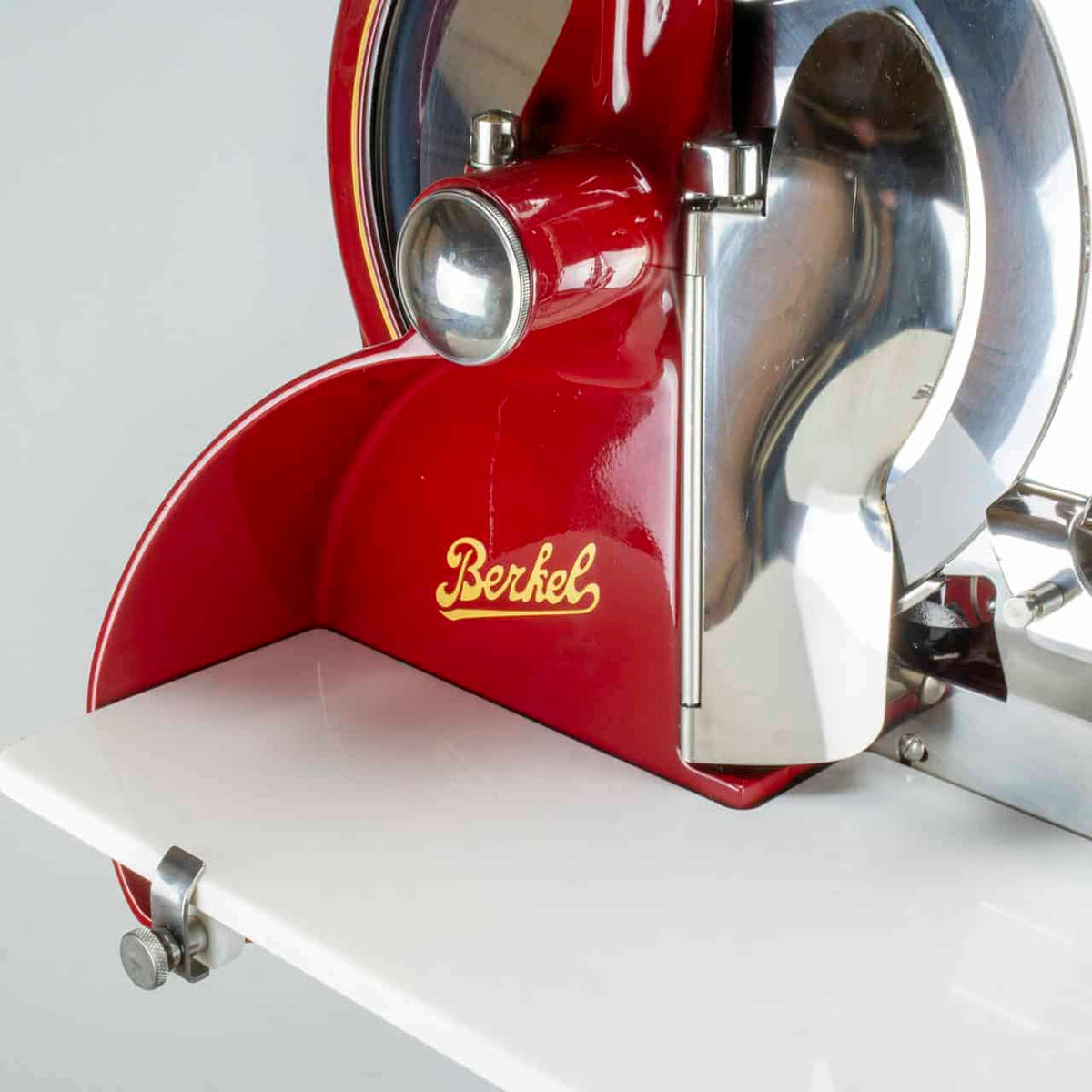 Eine Nahaufnahme einer roten Berkel Volano P15 Rot-Fleischschneidemaschine im Vintage-Stil mit Chromakzenten und glänzender Klinge. Das Wort „Berkel“ ist in gelber Schrift deutlich auf dem Hauptkörper zu sehen. Die Aufschnittmaschine Schwungrad + Standfuß + Aufschnittbrett ist auf einer weißen Plattform montiert, wobei ein Teil der Klemme sichtbar ist.