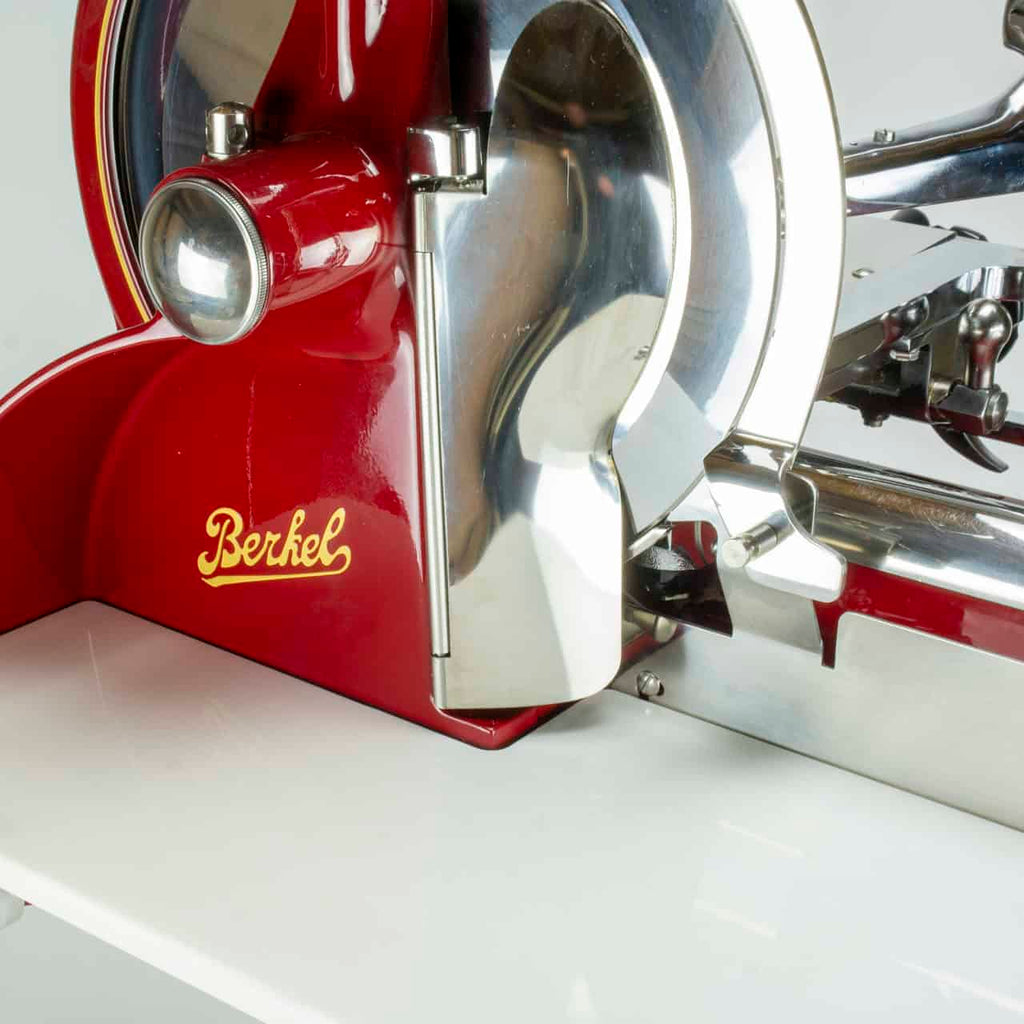Eine Nahaufnahme der Berkel Volano P15 Rot Aufschnittmaschine Schwungrad + Standfuß + Aufschnittbrett, die ihre glänzenden Metallteile und das deutlich sichtbare Berkel-Logo zeigt. Der Allesschneider steht auf einer sauberen, weißen Oberfläche.