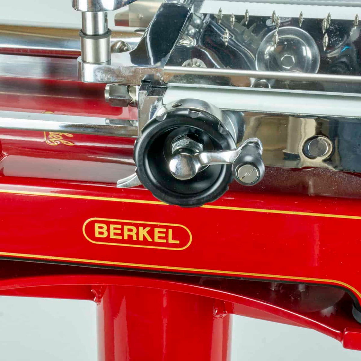 Nahaufnahme einer roten Berkel Volano P15 Rot Aufschnittmaschine mit Schwungrad + Standfuß + Aufschnittbrett. Die Aufschnittmaschine verfügt über glänzende Metallkomponenten, darunter ein scharfes Schneideblatt und Einstellräder. Der Herstellername „Berkel“ ist in gelber Schrift deutlich auf dem roten Sockel zu sehen.