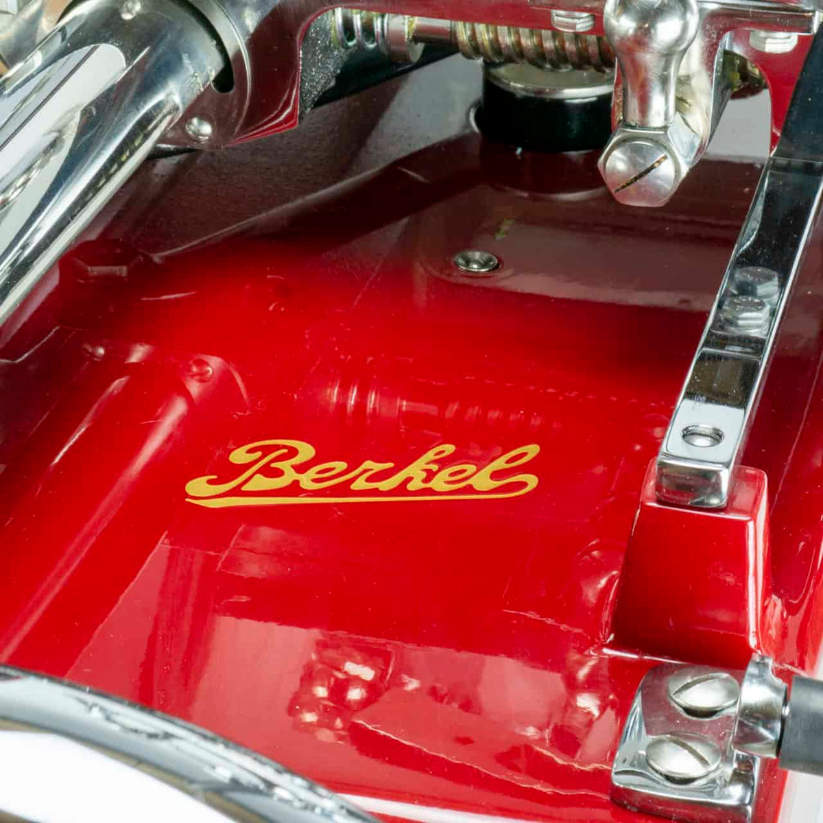 Eine Nahaufnahme einer Berkel Volano P15 Rot, Aufschnittmaschine Schwungrad + Standfuß + Aufschnittbrett, mit dem Markennamen „Berkel“ in gelber Schrift auf rotem Emaille. Das Bild fängt Details der Chromkomponenten und der glänzenden roten Oberfläche ein und betont das klassische Design und die für eine Schwungrad-Aufschnittmaschine charakteristische Handwerkskunst.