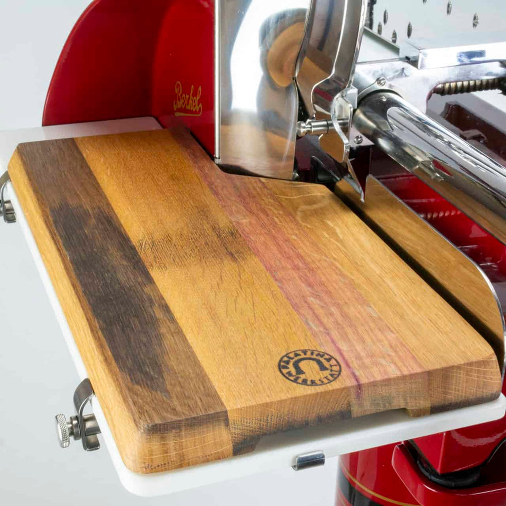 Eine Nahaufnahme zeigt ein Holzschneidebrett mit verschiedenen Holztönen auf einer Berkel Volano P15 Rot Aufschnittmaschine Schwungrad + Standfuß + Aufschnittbrett. Die Aufschnittmaschine ist metallisch mit einem markanten roten Schwungradabschnitt und das Brett weist ein rundes Markenlogo auf. Das Schneidebrett ist so positioniert, dass das geschnittene Fleisch direkt von der Klinge aufgefangen wird.