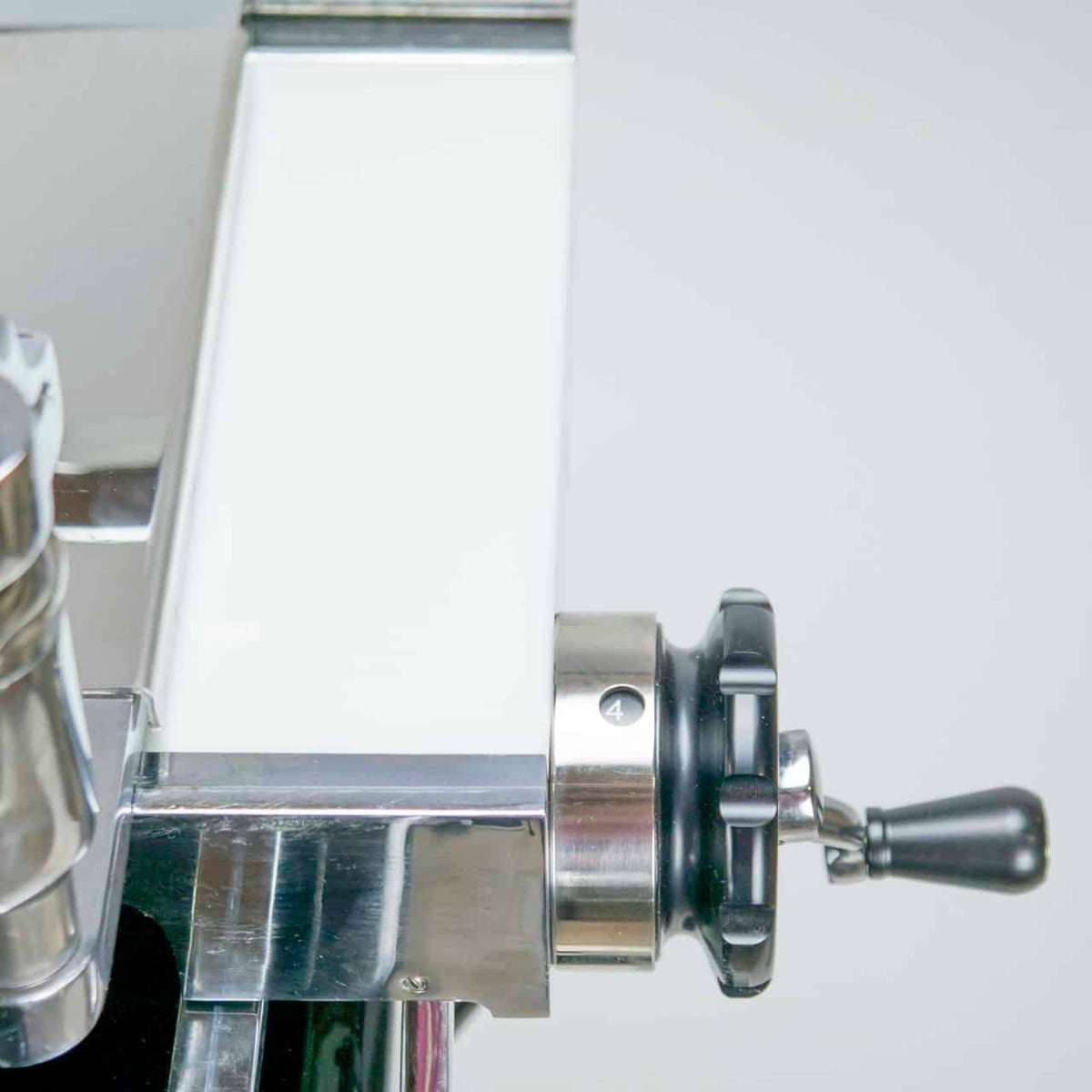 Nahaufnahme einer Berkel Volano P15 schwarz manuellen Aufschnittmaschine aus Edelstahl mit Schwungrad+Aufschnittbrett, mit einem Bedienknopf auf der rechten Seite. Der Knopf ist schwarz mit einem kleinen Griff und die Zahl „4“ ist auf seinem Rand sichtbar. Die Maschine hat neben dem Knopf eine glatte weiße Oberfläche.