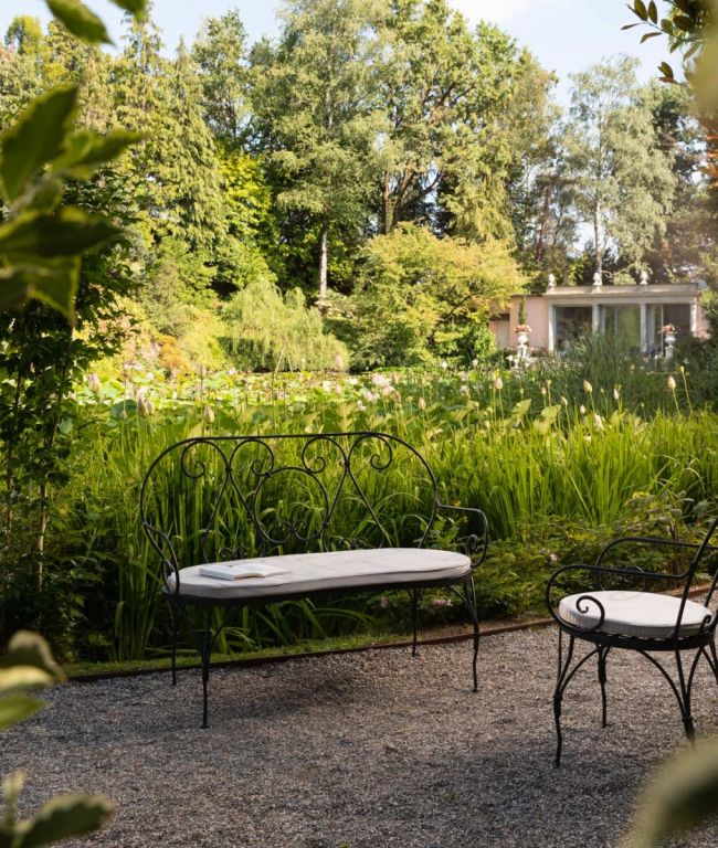 Eine anmutige Sitzbank Aurora aus Eisen mit weißen Kissen und einem Eisentisch steht auf einem Kiesweg in einem grünen Garten. Hohe grüne Pflanzen und Bäume umgeben die Umgebung, in der Ferne lugt ein kleines Gebäude hervor. Sonnenlicht strömt durch die Blätter und schafft eine Atmosphäre der Ruhe.