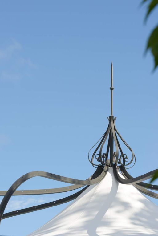 Eine hohe, dekorative Metallspitze ziert die Spitze eines weißen Pavillon Solaire vor einem klaren blauen Himmel. Die aufwendige Metallarbeit zeichnet sich durch elegante, geschwungene Details und eine schlanke, spitze Spitze aus. Auf der rechten Seite des Bildes wirft ein Teil eines Astes mit grünen Blättern einen bezaubernden Schatten auf die Solaire-Struktur.