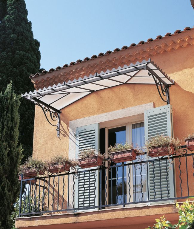 Ein malerisches orangefarbenes Stuckgebäude mit hellblauen Fensterläden und einem dekorativen schmiedeeisernen Geländer, das mit Blumenkästen geschmückt ist. Die Tür zum Balkon wird durch das Vordach mit Flachdach Dafne aufgewertet, das einen dreieckigen Metallrahmen und ein lichtdurchlässiges Dach aufweist. Das Ganze steht unter hohen Bäumen, die dem Gebäude seinen Charme verleihen.