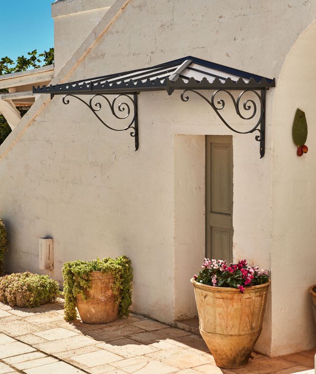 Eine malerische Szene im Freien zeigt eine rustikale weiße Gipswand mit einer kleinen grünen Tür unter einem dekorativen schwarzen schmiedeeisernen Vordach mit Flachdach Dafne. Topfpflanzen und Sträucher sind um den Eingang herum platziert und schaffen eine gemütliche und einladende Atmosphäre.