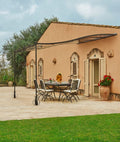 Eine geräumige Terrasse neben einem rustikalen Stuckhaus verfügt über einen runden Esstisch mit sechs Stühlen unter einer Anbaupergola Tibisco mit Überdachung aus Polykarbonat. Topfpflanzen schmücken den Bereich und das Haus hat mehrere Fenster und Türen mit Fensterläden. Im Vordergrund erstreckt sich ein üppiger grüner Rasen.