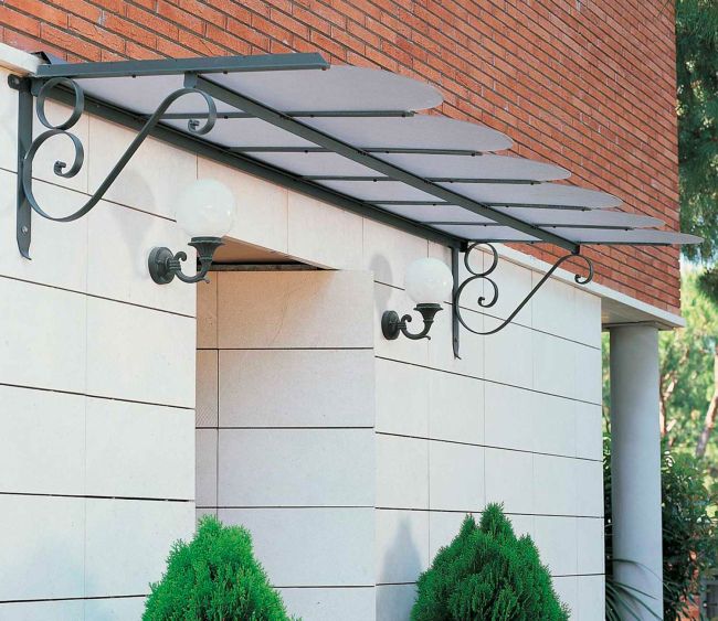Ein moderner Gebäudeeingang mit einem Vordach mit Flachdach Dafne, das von verzierten Konsolen getragen wird. Zwei kugelförmige Wandleuchten sind an der Wand unter der Markise befestigt. Die Wand besteht aus einer Mischung aus rotem Backstein und hellem Stein und im Vordergrund sind grüne Sträucher zu sehen.