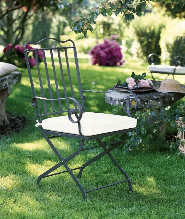 Ein schwarzer klappbarer Gartenstuhl „Stuhl Toscana“ mit weißem Kissen steht auf einer Rasenfläche. In der Nähe steht ein Steintisch mit einem Korb, einem Hut und einer Brille. Die Toskana-Szene ist von üppigem Grün und Blumen umgeben. Im Hintergrund ist ein weiterer Stuhl teilweise sichtbar.