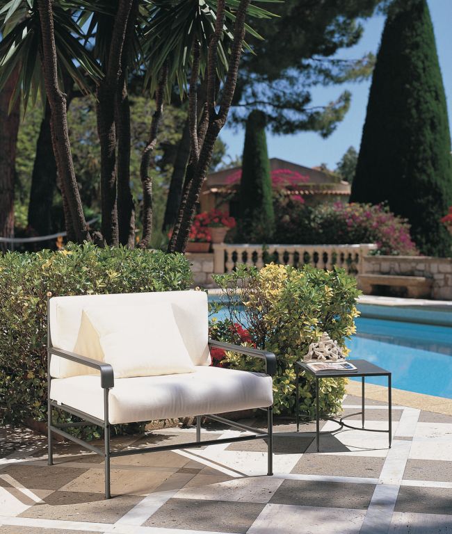 Eine ruhige Außenumgebung besteht aus einem gepolsterten graphitgrauen Sessel Toscana aus Eisen neben einem kleinen Beistelltisch mit einem dekorativen Objekt auf einer gefliesten Terrasse neben einem Pool. Im Hintergrund sieht man üppiges Grün, hohe Bäume, bunte Blumen und einen Blick auf ein Haus mit Eisenzaun, das den Charme der Toskana heraufbeschwört.
