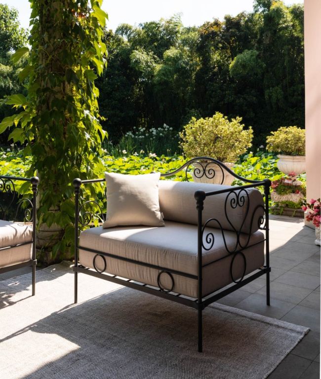 Ein schmiedeeiserner „Sessel Aurora aus verzinktem Eisen“ mit beigen Kissen steht auf einem grauen Teppich auf einer Terrasse. Üppiges, grünes Laub und Bäume bilden eine malerische Kulisse. Eine Topfpflanze und ein Blumenarrangement schmücken den Terrassenbereich und verstärken die ruhige Atmosphäre im Licht der Aurora-Dämmerung.