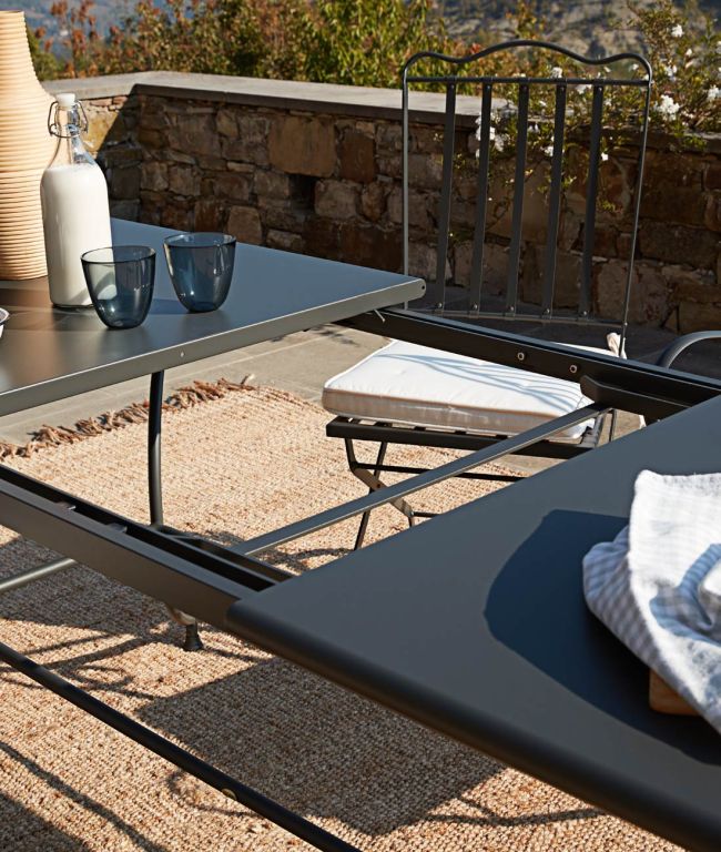 Ein moderner Essbereich im Freien verfügt über den Tisch Toscana rechteckig 180 cm, einen rechteckigen schwarzen Metalltisch mit zwei blauen Gläsern, einer Flasche und einer Vase. Zwei schwarze Metallstühle mit weißen Kissen stehen um den Tisch herum. Das von der Toskana inspirierte Arrangement steht auf einer Steinterrasse mit einem Naturfaserteppich vor einer charmanten Steinmauer.