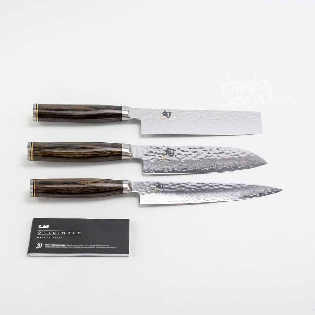 Drei hochwertige Küchenmesser aus dem Exklusiven Messerblock-Set der KAI Shun Tim Mälzer Damastmesser-Kollektion mit Holzgriffen werden auf einer weißen Oberfläche präsentiert. Jede Klinge weist eine einzigartige gehämmerte Textur auf. Eine Visitenkarte mit der Aufschrift „Kai Originals“ liegt neben den Messern und ergibt ein elegantes Tableau des Messerblock-Sets mit einem hochwertigen Magnet-Messerblock von Böker.