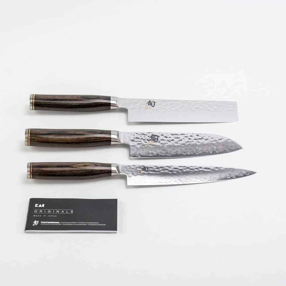 Drei hochwertige Küchenmesser aus dem Exklusiven Messerblock-Set der KAI Shun Tim Mälzer Damastmesser-Kollektion mit Holzgriffen werden auf einer weißen Oberfläche präsentiert. Jede Klinge weist eine einzigartige gehämmerte Textur auf. Eine Visitenkarte mit der Aufschrift „Kai Originals“ liegt neben den Messern und ergibt ein elegantes Tableau des Messerblock-Sets mit einem hochwertigen Magnet-Messerblock von Böker.