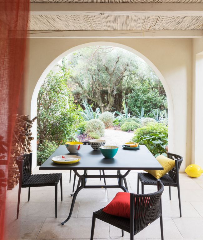 Eine Terrasse mit einem quadratischen Tisch Toscana, einem schwarzen Tisch für vier Personen unter einem überdachten Torbogen. Auf dem Tisch sind bunte Schüsseln und Teller angeordnet, und schwarze Stühle mit roten und gelben Kissen umgeben ihn. Im Hintergrund ist ein üppiger Toscana-Garten mit verschiedenen Grünpflanzen zu sehen.