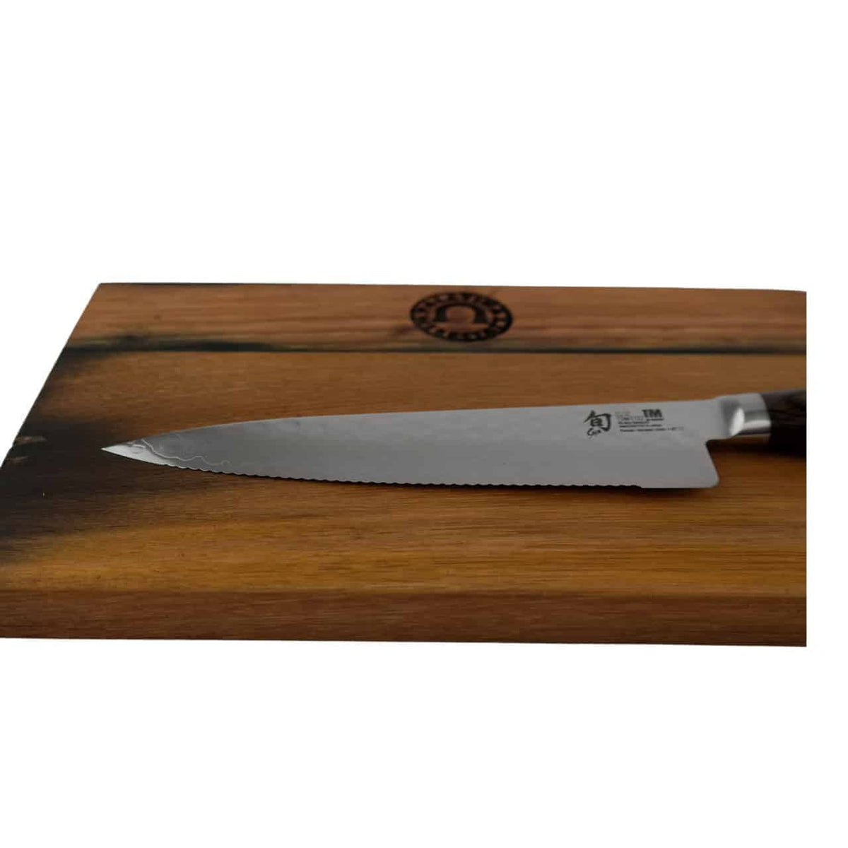 Ein KAI Shun Premier Tim Mälzer TDM-1722 Allzweckmesser mit gezahnter 16,5 cm Klinge und dunklem Griff liegt auf einem Fassholz Schneidebrett 25x15 cm. Das Schneidebrett ist im oberen Mittelteil mit einem Markenlogo versehen.