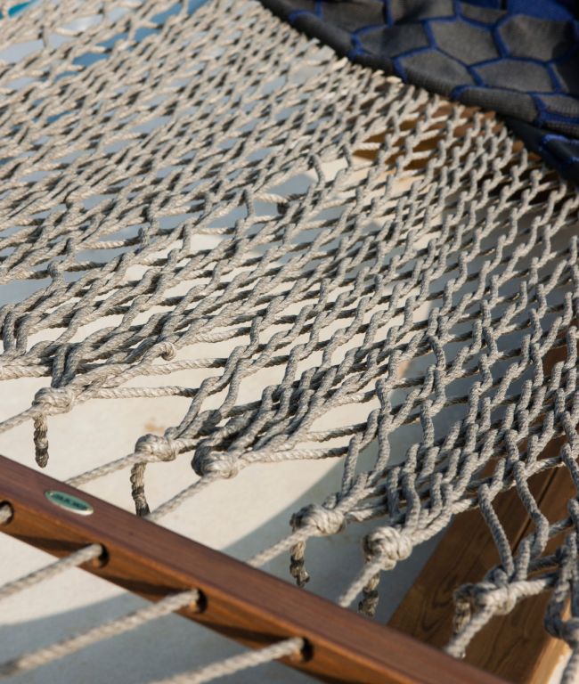 Nahaufnahme der Hängematte komplett mit Netzwerk aus geknotetem Seil, befestigt an einem Holzrahmen. Die Hängematte ist leicht beschattet und das Sonnenlicht hebt das komplizierte Muster der Seile hervor. Im Hintergrund ist ein marineblaues Tuch zu sehen, das die Hängematte teilweise bedeckt und ein fast nahtloses Netzwerk bildet.