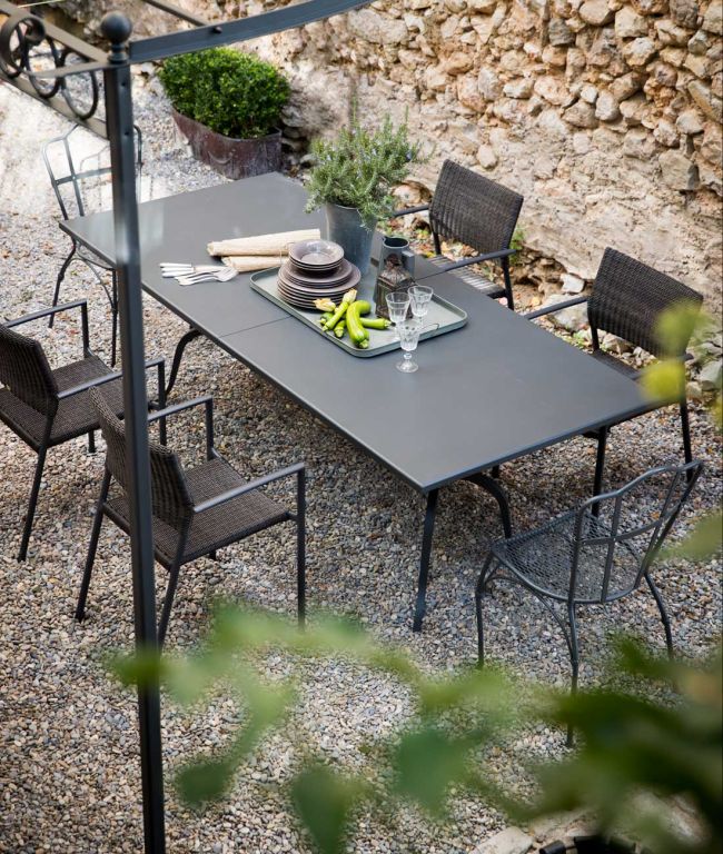 Ein moderner Esstisch für sechs Personen mit schwarzen Stühlen, aufgestellt auf einer Kiesfläche neben einer Steinmauer. Der rechteckige Tisch Toscana (220 cm) ist mit einem Tablett ausgestattet, auf dem Teller, Besteck, Gläser, Bananen, Brot und Topfkräuter liegen. Topfpflanzen umgeben den ruhigen, von der Toskana inspirierten Bereich.