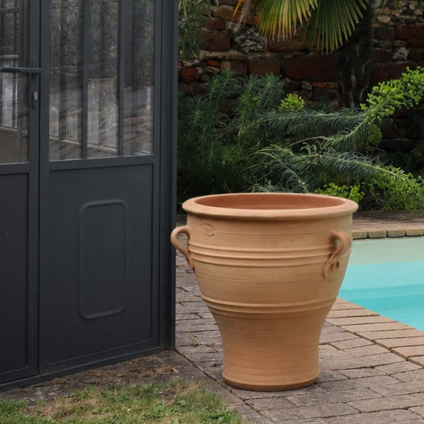 Das Terrakotta-Pflanzgefäß "Rosa", H=40 cm (Kopie), mit zwei Henkeln, steht auf einer Steinterrasse durch eine schwarze Glastür mit Metallrahmen in der Nähe des Pools, umgeben von Grünpflanzen und einer Steinmauer - ideal für jeden Gartenbereich.