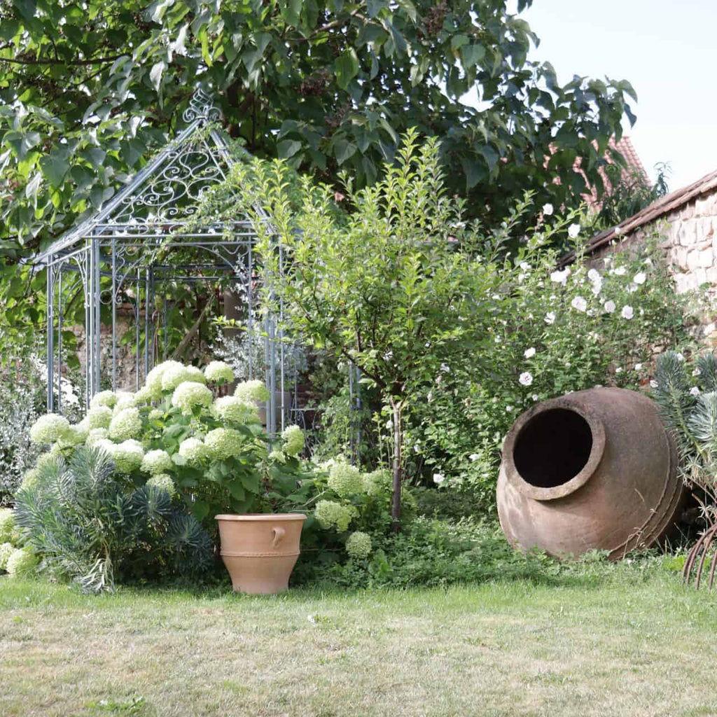 Ein üppiger Garten besteht aus einem großen Terrakotta-Pflanzkübel, einem kleineren Terrakotta-Topf, blühenden weißen Hortensien und einem Baum. Eine dekorative Pavillonstruktur aus Metall steht inmitten des Grüns. Die Kulisse besteht aus einer Steinmauer, die teilweise mit Grün bedeckt und mit Pflanzen des Terrakotta-Pflanzkübels „Rosa 2“, H=40 cm, übersät ist.