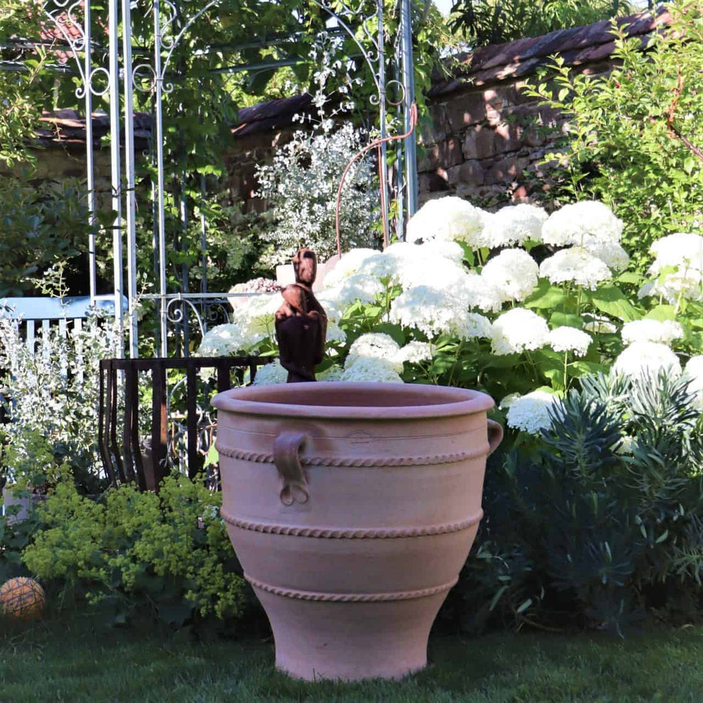 Ein großer, verzierter Terrakotta-Pflanzkübel „Rosa 2“ mit 60 cm Höhe und dekorativem Griff steht in einem üppigen Garten. Weiße Hortensien und verschiedene Grünpflanzen umgeben ihn. Im Hintergrund sind ein Eisenspalier und eine alte Steinmauer zu sehen, die dem Garten Charme verleihen.