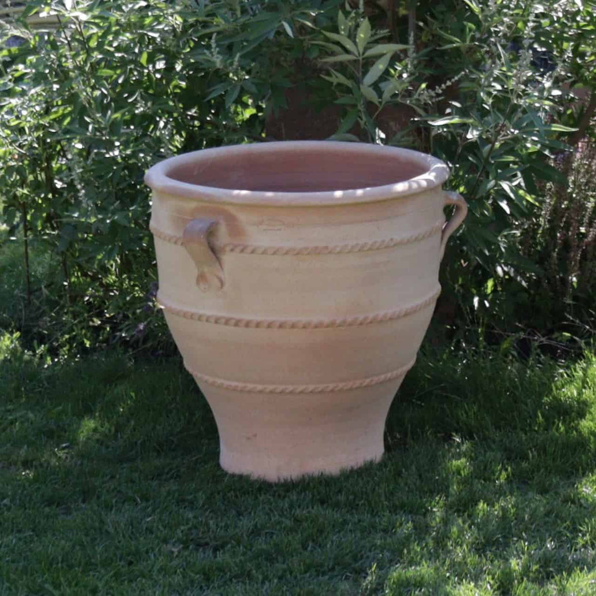 Ein großer, leerer Terrakotta-Pflanzkübel „Rosa 2“, 60 cm hoch, mit zwei Griffen und dekorativen seilartigen Mustern, die ihn umgeben, steht auf einem gepflegten Rasen. Im Hintergrund sind grüne Blätter und Pflanzen zu sehen, die eine natürliche und üppige Umgebung bilden, die seine Eleganz perfekt ergänzt.