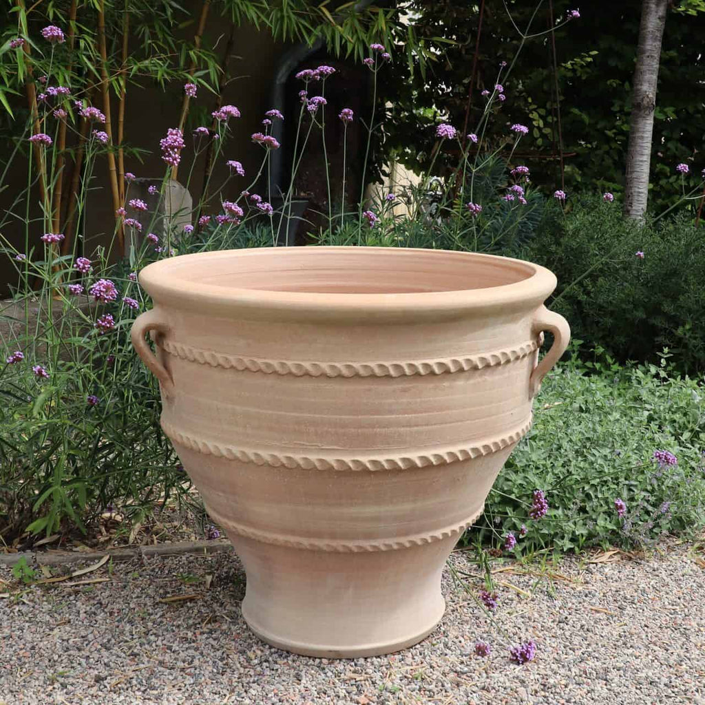 Der große Terrakotta-Pflanzkübel, Pflanzgefäß „Rosa 2“ H=70cm mit dekorativen horizontalen Rillen und zwei Griffen steht in einem Kiesbereich. Hohe violette Blumen und grünes Laub füllen den Hintergrund und schaffen eine natürliche Gartenkulisse.
