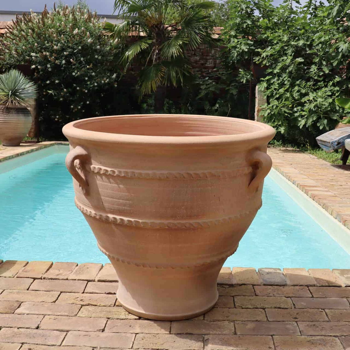 Ein großer, leerer Pflanzkübel aus Terrakotta, genauer gesagt das Pflanzgefäß „Rosa 2“ mit einer Höhe von 70 cm, geschmückt mit dekorativen Griffen, steht auf einer mit Steinfliesen ausgelegten Terrasse neben einem klaren Swimmingpool. Die Szene ist von Grün umgeben, darunter Palmen und Büsche, die ein entspanntes, tropisches Ambiente schaffen.