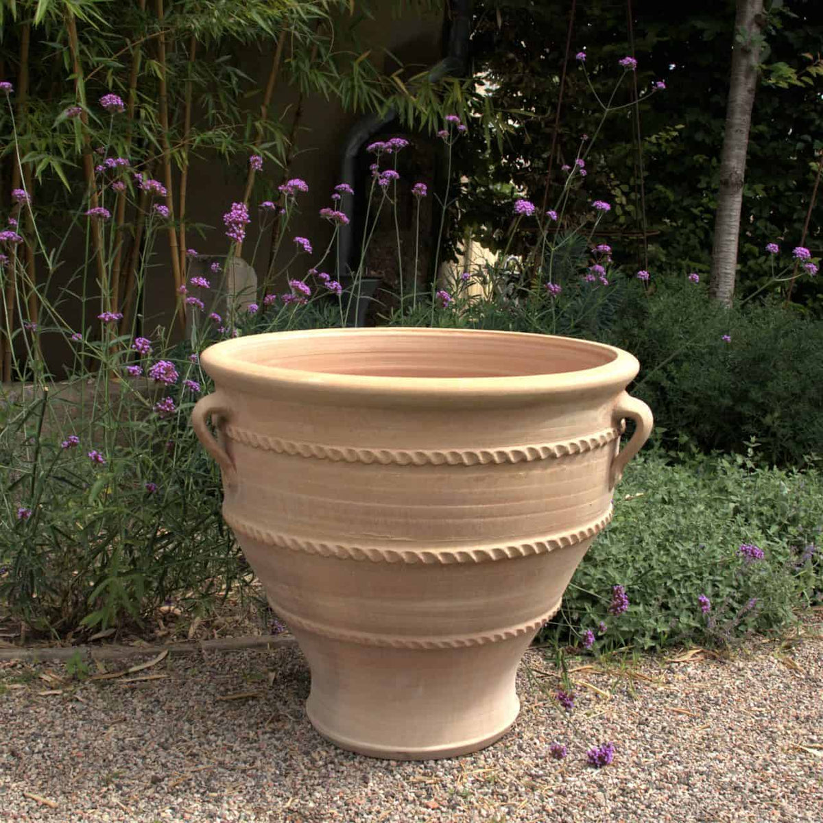 Ein großer Terrakotta-Pflanzkübel, Pflanzgefäß „Rosa 2“ H=70cm mit zwei seitlichen Griffen und horizontalen Rillen steht auf einer Kiesfläche. Im Hintergrund sind violette Blumen und grünes Laub zu sehen, die eine ruhige Gartenkulisse schaffen, die an Rosa 2 erinnert.