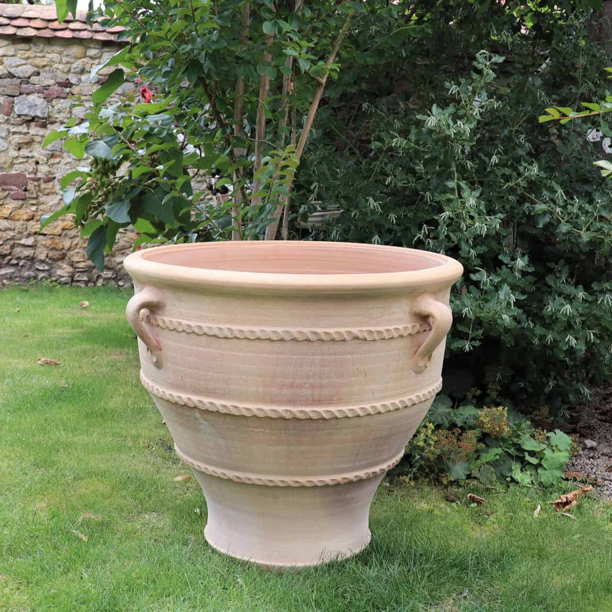Ein großer Terrakotta Pflanzkübel, Pflanzgefäß "Rosa 2" H=70cm in Beige mit zwei dekorativen gedrehten Seilmustern und Henkeln steht auf einem gepflegten Rasen. Im Hintergrund sieht man Grünpflanzen, darunter einen Baum und einige Sträucher, sowie eine Steinmauer.