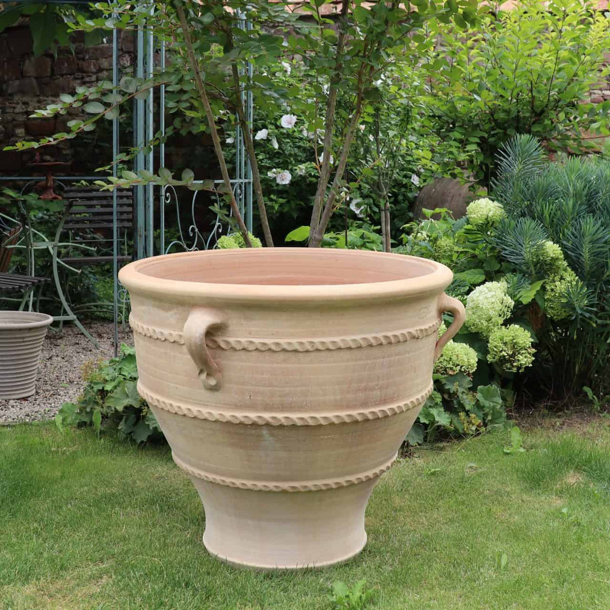 Ein großer Terrakotta-Pflanzkübel „Rosa 2“ mit dekorativen gemusterten Bändern und zwei Griffen steht auf einer Rasenfläche in einem üppigen Garten. Um das Pflanzgefäß „Rosa 2“ H=70cm herum stehen verschiedene Grünpflanzen und Sträucher, teilweise beschattet von den Ästen eines Baumes. Im Hintergrund ist ein Metallstuhl zu sehen.