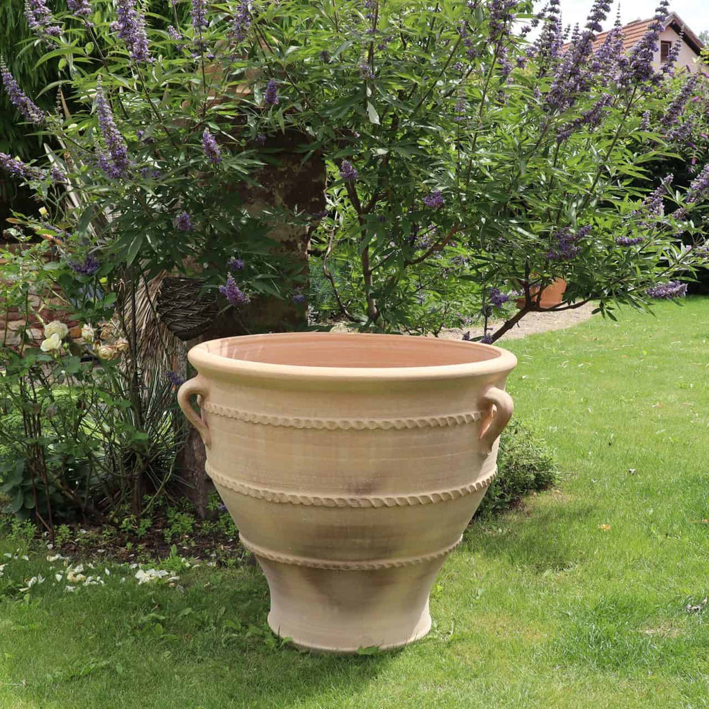 Ein großer Terrakotta-Pflanzkübel, Pflanzgefäß „Rosa 2“ H=70cm mit dekorativen Griffen steht auf einem gepflegten grünen Rasen. Er ist vor einem üppig blühenden Busch mit violetten Blüten platziert und trägt zu einer ruhigen Gartenumgebung bei.