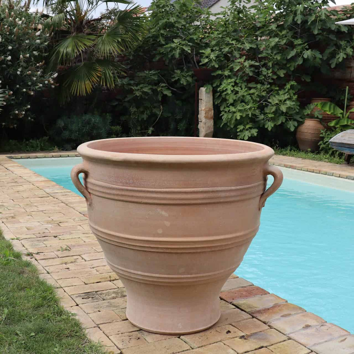 Ein großer, leerer Terrakotta-Pflanzkübel „Rosa“ H=70cm mit zwei Griffen steht auf dem Rand eines rechteckigen Swimmingpools. Der Pool ist von einem gepflasterten Bereich und üppigem Grün, darunter Bäumen und Pflanzen, umgeben, was eine ruhige Außenumgebung schafft. Der Terrakotta-Pflanzkübel „Rosa“ hat eine beeindruckende Höhe von 70cm.