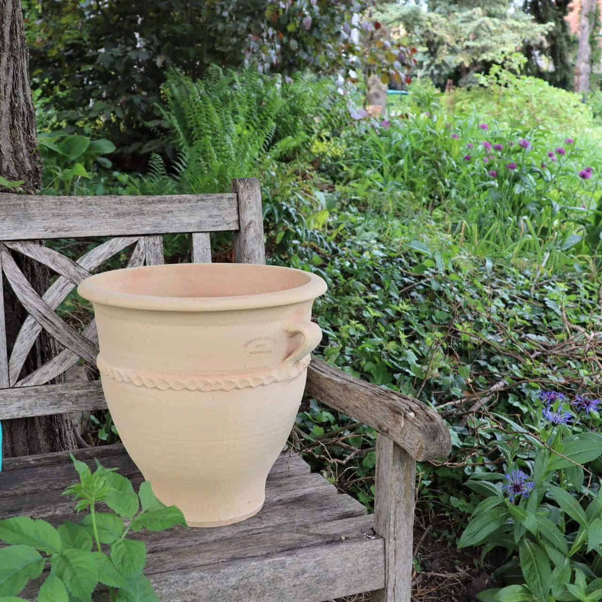 Ein großer, beiger Tontopf mit dekorativen Rillen auf der Oberfläche steht auf einer verwitterten Holzbank in einem üppigen Garten. Um die Bank herum stehen verschiedene Grünpflanzen und leuchtende Blumen, die eine ruhige und natürliche Atmosphäre schaffen. Der Terrakotta-Pflanzkübel „Rosa 2“ (H=35 cm) verleiht der Szene einen erdigen Charme.