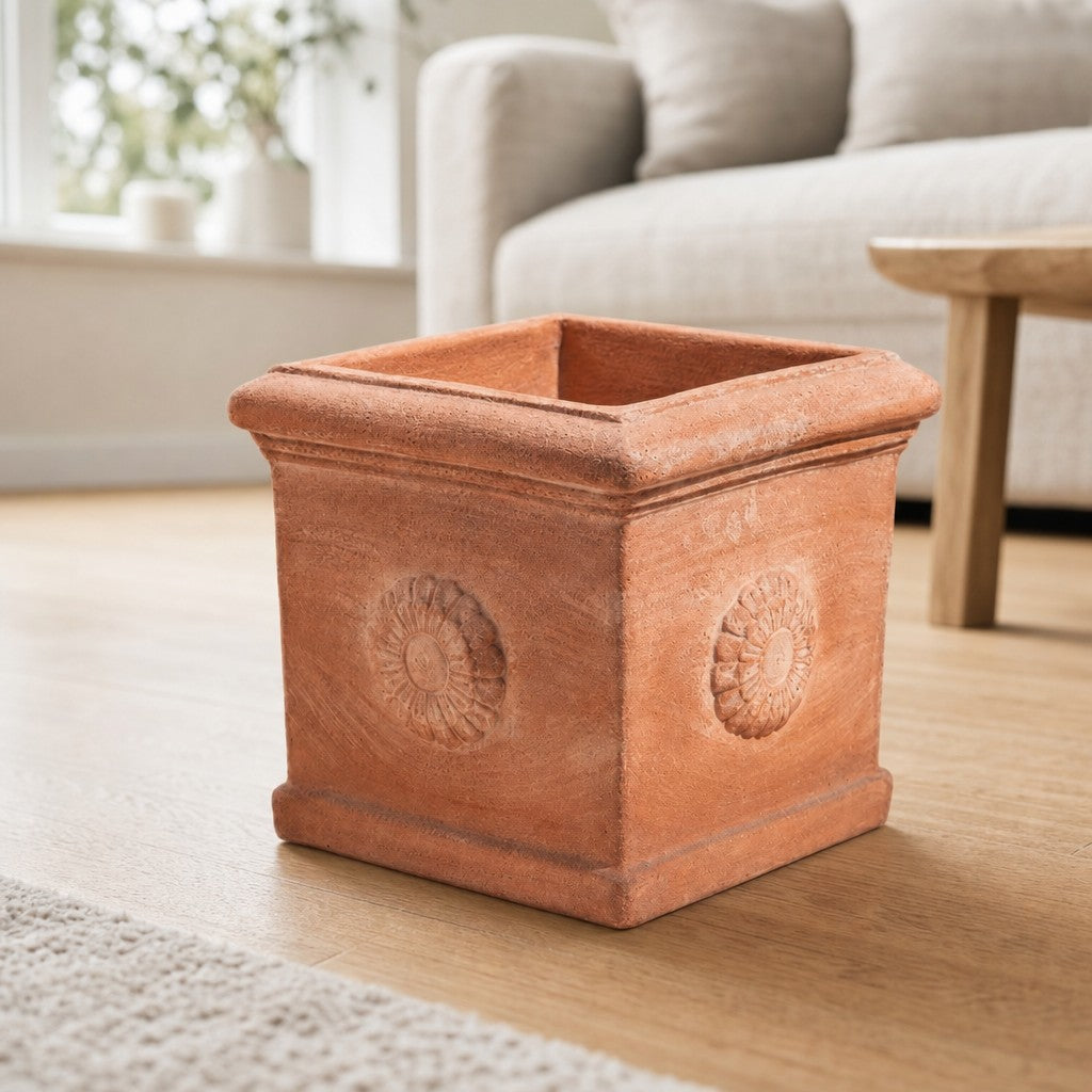 Ein quadratischer Terrakotta-Kübel „Rosone“ mit geprägten Blumenmotiven steht auf einem Holzboden in einem hellen Wohnzimmer neben einem Sofa und einem Couchtisch.